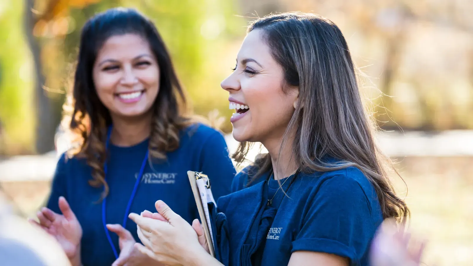 Two SYNERGY HomeCare team members in branded shirts, talking and smiling